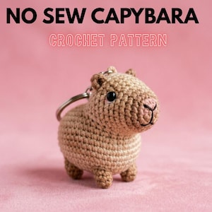 May include: A crocheted capybara keychain in tan and brown, with a black eye and pink nose. The keychain is attached to a silver ring. Text at the top reads "NO SEW CAPYBARA" and "CROCHET PATTERN".