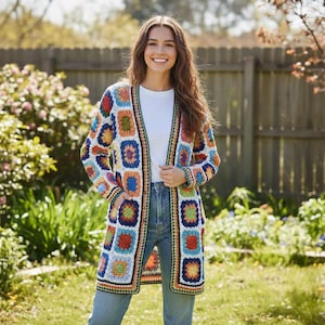 Könnte beinhalten: Ein langer, offener Häkelcardigan mit einem farbenfrohen Patchwork-Design. Der Cardigan zeigt quadratische Muster in Blau, Orange, Grün und Gelb, umrandet in Schwarz und Weiß. Das Kleidungsstück wird über einem weißen Oberteil und einer blauen Jeans getragen.