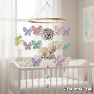 Könnte beinhalten: Handgefertigtes Baby-Mobile mit einem schlafenden gehäkelten Baby, umgeben von pastellfarbenen Schmetterlingen und floralen Akzenten. Das Mobile enthält einen Holzring und hängende Kugeln in sanften Farben, wodurch eine skurrile Kinderzimmerdekoration entsteht.