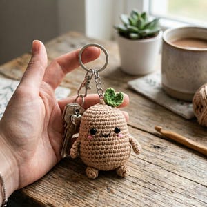 Puede incluir: Un llavero de ganchillo con un personaje sonriente de color marrón, parecido a una verdura de raíz, con una hoja verde en la parte superior. El llavero está unido a un llavero plateado con llaves.