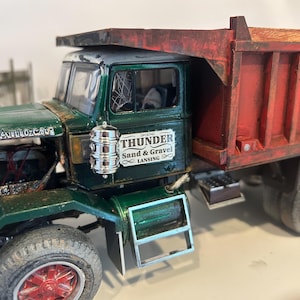 May include: A detailed model of a vintage green and red dump truck. The truck features the text "THUNDER Sand & Gravel LANSING" on the side. The truck has a weathered appearance, with visible rust and wear.