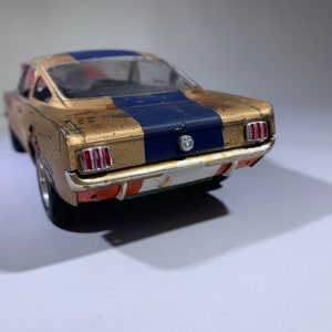 May include: A gold and blue model car with a weathered finish. The car has a blue racing stripe down the center of the hood and roof. The car is a classic American muscle car.