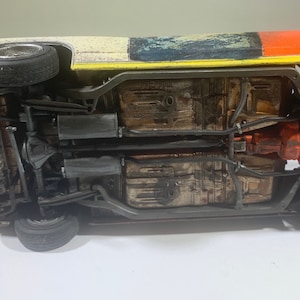 May include: Underside view of a model car with a rusty brown undercarriage, black exhaust pipes, and a red engine. The car is painted white with blue and yellow accents.