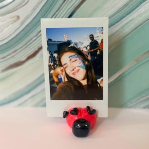 Może przedstawiać: Zdjęcie Polaroid trzymane przez czerwony uchwyt na zdjęcia w kształcie serca. Zdjęcie przedstawia osobę z malunkiem na twarzy. Uchwyt ma czarne akcenty i znajduje się na białej powierzchni.