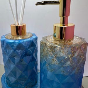 Könnte beinhalten: Blaues Badezimmer-Set aus Kunstharz mit geometrischem Design. Das Set enthält einen Seifenspender mit goldener Pumpe und einen Diffusor mit weißen Stäbchen. Goldflocken sind in das Harz eingebettet, und das Set steht auf einem passenden Tablett.