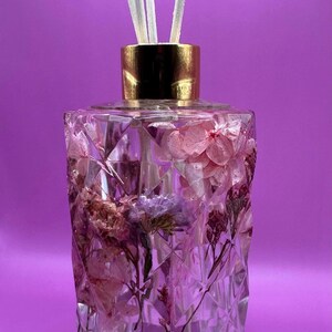 Könnte beinhalten: Eine klare, facettierte Glas-Diffusorflasche mit goldenem Deckel, gefüllt mit getrockneten Blumen in Rosa-, Lila- und Brauntönen. Weiße Schilfrohre ragen aus der Oberseite heraus. Der Hintergrund ist einfarbig lila.
