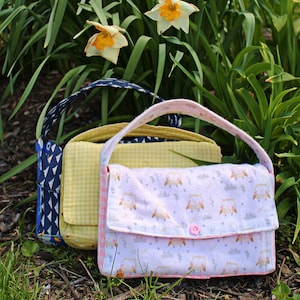 Op de afbeelding: Drie handgemaakte stoffen handtassen met handvatten. De voorste tas is lichtroze met een uilenpatroon en een roze knoopsluiting. De middelste tas is geel geruit en de achterste tas is marineblauw met een wit geometrisch patroon.