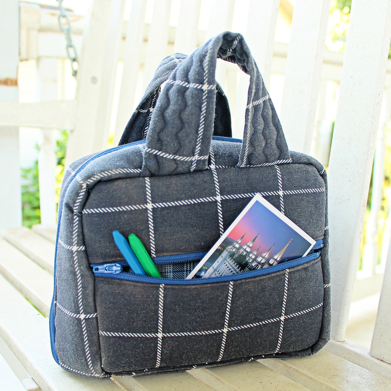 Puede incluir: Una peque&ntilde;a bolsa de tela azul y blanca a cuadros con un bolsillo con cremallera y un asa. La bolsa tiene dos l&aacute;pices de colores y una postal con una imagen de un edificio dentro del bolsillo.