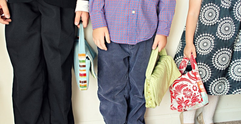 May include: Three children holding colorful tote bags. The bags have various patterns, including stripes, floral, and geometric designs. The children are wearing different outfits, including a dress, pants, and a button-down shirt.