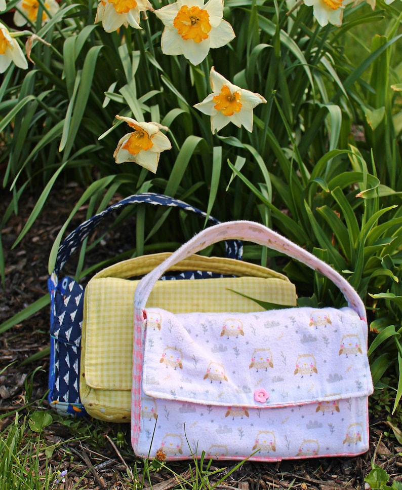 May include: Three handmade fabric purses with handles. The front purse is light pink with an owl pattern and a pink button closure. The middle purse is yellow gingham, and the back purse is navy blue with a white geometric pattern.