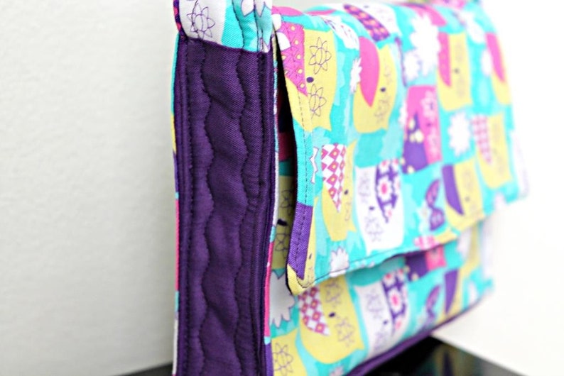 May include: A quilted clutch purse with a flap closure. The purse features a vibrant pattern of colorful shapes on a turquoise background. The edges are trimmed with a deep purple fabric, and the interior is also purple.
