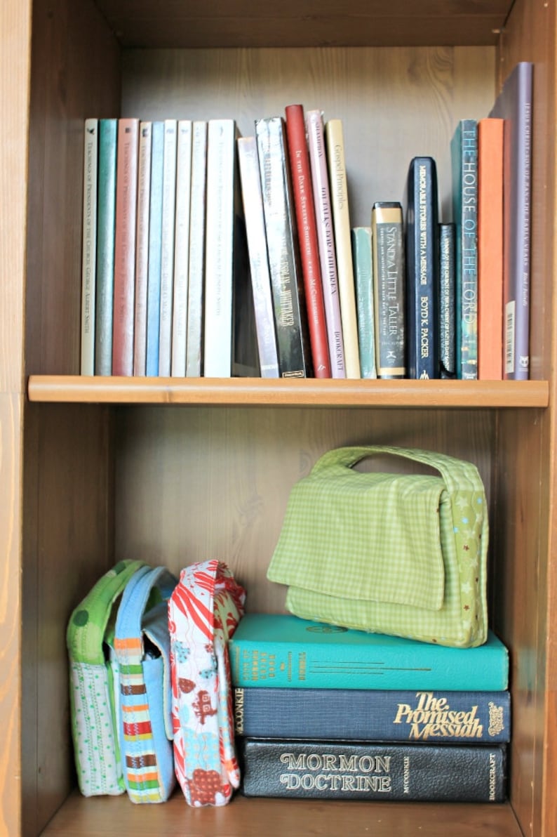 May include: A wooden bookshelf filled with books and fabric bags. The top shelf holds a variety of books with colorful spines. The bottom shelf displays a stack of books and several patterned bags, one green with a handle.