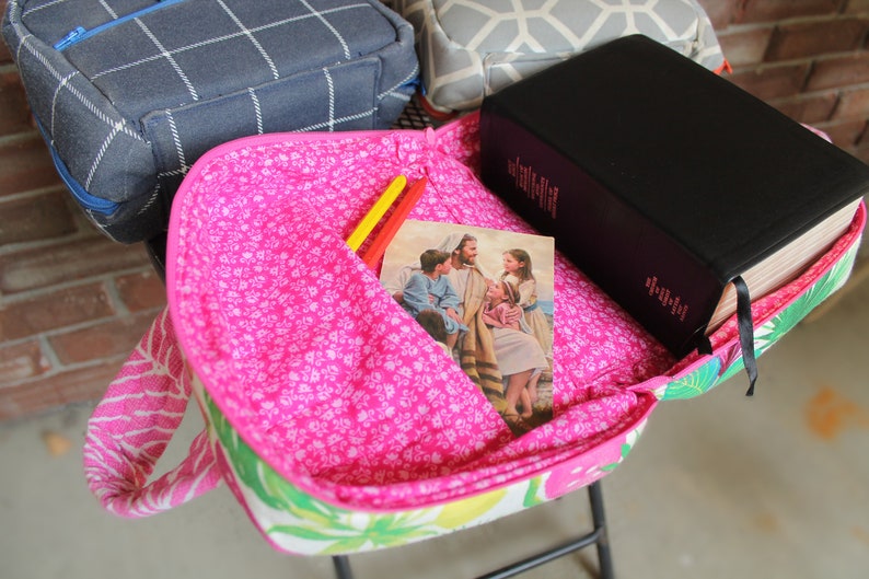 Puede incluir: Una bolsa de tela rosa y verde con un libro negro, una imagen de Jes&uacute;s y ni&ntilde;os, y dos l&aacute;pices amarillos y naranjas en su interior.