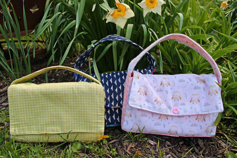 May include: Three handmade fabric handbags in a garden setting. One is light green with a gingham pattern, another is navy blue with a white geometric design, and the third is pink with an owl print.