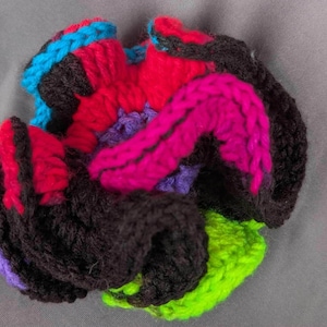 May include: A handmade, multi-colored crocheted flower. The flower features vibrant hues of black, red, pink, purple, blue, and green. The yarn is tightly woven, creating a textured and dimensional effect. This unique piece could be used as a decorative accent or embellishment.