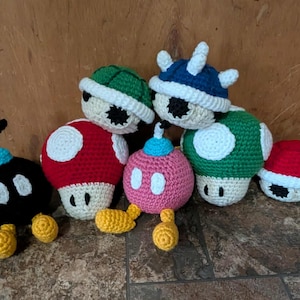 Op de afbeelding: Een verzameling handgemaakte haakfiguren geïnspireerd op een videogame. De figuren omvatten een zwarte bom met een blauwe lont, rode en groene paddenstoelen, een roze bom en blauwe, groene en rode schildpadden, allemaal met witte accenten.