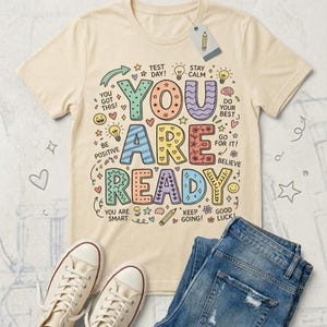 May include: Cream-colored t-shirt with the words "YOU ARE READY" in colorful, whimsical lettering. The shirt has encouraging phrases and doodles around the main text, with a pair of blue jeans and white sneakers in the image.