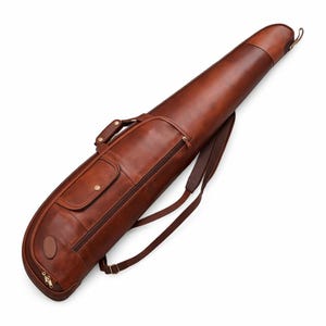 Könnte beinhalten: Braune Leder-Gewehrhülle mit Schulterriemen und Griff. Die Hülle hat eine Reißverschlusstasche, eine kleine Klapptasche und einen messingfarbenen Reißverschluss. Konzipiert für den Transport einer Langwaffe.
