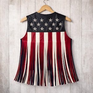 Back view of American flag fringe leather vest displayed on a wooden hanger. Patriotic women’s vest with black leather top, silver star details, red white blue stripes, and long western fringe. Handmade USA inspired leather vest for 4th of July outfits.
