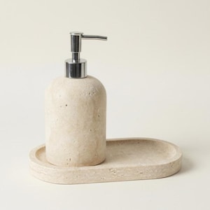 May include: A beige stone soap dispenser with a silver pump, on a matching oval tray. The dispenser has a rounded shape and a textured surface. The set is designed for bathroom use.