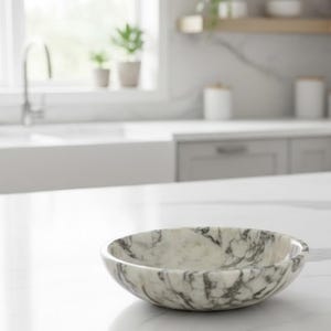 Puede incluir: Un cuenco de m&aacute;rmol con un patr&oacute;n gris y blanco se encuentra sobre una encimera blanca. El cuenco tiene forma redondeada y una superficie lisa. El entorno de la cocina incluye una ventana, un fregadero y armarios.