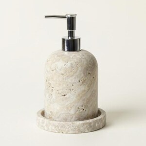 May include: A beige stone soap dispenser with a silver pump. The dispenser has a rounded shape and sits on a matching circular base. The stone has a natural, textured appearance.