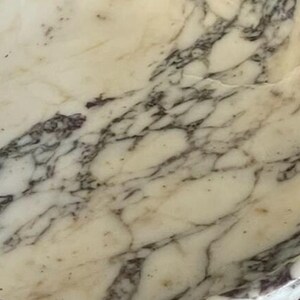 Puede incluir: Primer plano de una superficie de m&aacute;rmol crema con vetas grises oscuras. El patr&oacute;n de piedra natural presenta l&iacute;neas y formas irregulares, creando una textura &uacute;nica. Ideal para la decoraci&oacute;n del hogar y proyectos de dise&ntilde;o.