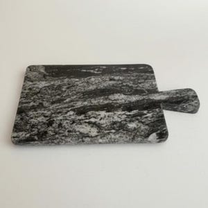 May include: A rectangular cutting board with a handle, crafted from gray and black marble. The board features a natural, swirled pattern, and is designed for food preparation and serving. The handle is rounded for easy grip.