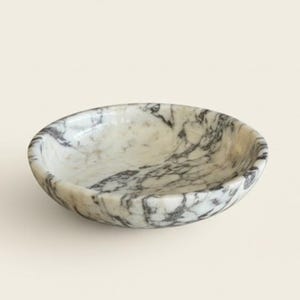 Puede incluir: Un cuenco peque&ntilde;o y redondo de m&aacute;rmol con base blanca y vetas grises. El cuenco tiene una superficie lisa y pulida y una profundidad baja. Este cuenco decorativo es adecuado para guardar objetos peque&ntilde;os.