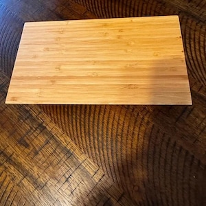 Könnte beinhalten: Ein rechteckiges Schneidebrett aus Bambus in hellbrauner Farbe. Das Brett hat eine glatte Oberfläche und besteht aus mehreren Bambusstreifen. Das Schneidebrett liegt auf einer dunkelbraunen Holzoberfläche.
