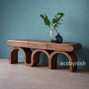 Op de afbeelding: Houten bank met een unieke gebogen basis en een gladde, rechthoekige bovenkant. De bank is gemaakt van warm getint hout en heeft een modern design. Twee vazen met groene bladeren staan op de bank. Het woord "ecobynish" staat rechtsonder.