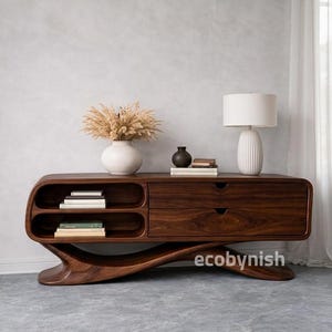 Puede incluir: Consola de TV moderna de madera oscura con base curva y estanterías integradas. La consola tiene dos cajones y una superficie superior con objetos decorativos, incluyendo una lámpara y un jarrón blancos. El texto "ecobynish" es visible.