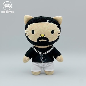 May include: A soft toy cat with a black beard, mustache, and hairstyle. It is dressed in a black t-shirt with an owl design, white shorts, and a silver chain. A heart shape is on its head. The toy is a novelty item.