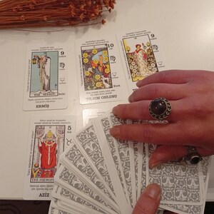 May include: A collection of tarot cards with various illustrations and Turkish text. The cards are spread out on a white surface, with a hand holding a deck and wearing a dark stone ring. Dried brown plant stems are in the background.