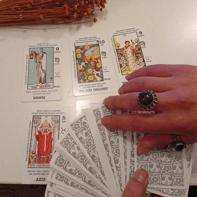 May include: A collection of tarot cards with various illustrations and Turkish text. The cards are spread out on a white surface, with a hand holding a deck and wearing a dark stone ring. Dried brown plant stems are in the background.