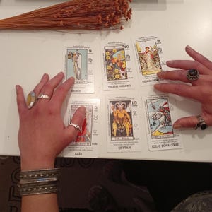 May include: A tarot card reading with several cards laid out, featuring illustrations and text. The cards are being handled by hands adorned with rings and bracelets. Dried flowers are in the background.