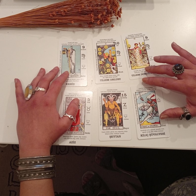 May include: A tarot card reading with several cards laid out, featuring illustrations and text. The cards are being handled by hands adorned with rings and bracelets. Dried flowers are in the background.