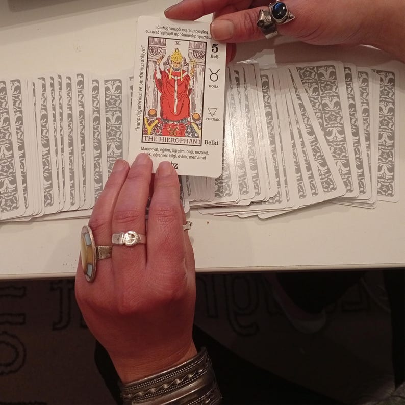 May include: A tarot card with the image of The Hierophant, card number 5, held over a deck of cards. The Hierophant is depicted in red robes with a golden crown. The card has text in a foreign language.