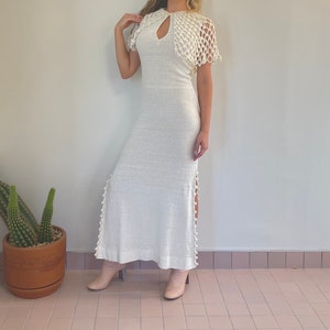 May include: A white crochet maxi dress with a keyhole neckline and a crochet cape. The dress has a side slit and is adorned with white pom-poms.