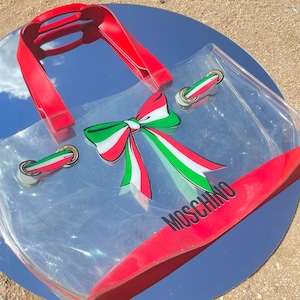 May include: Clear plastic tote bag with red trim and a red, white, and green bow. The bag has gold hardware and the word "MOSCHINO" printed on the bottom.