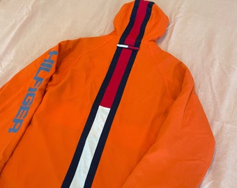 May include: Orange hooded jacket with a white, red, and blue stripe down the back. The jacket has "HILFIGER" written on the left sleeve and on the right cuff.