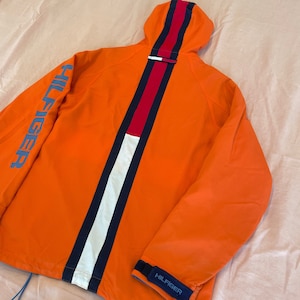May include: Orange hooded jacket with a white, red, and blue stripe down the back. The jacket has "HILFIGER" written on the left sleeve and on the right cuff.