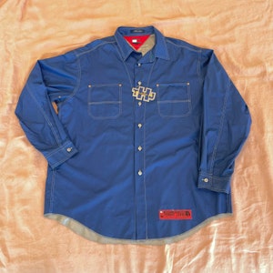 May include: A blue long-sleeve button-down shirt with white stitching and a Tommy Jeans logo patch. The shirt has two chest pockets with white stitching and a metal Tommy Jeans logo charm.