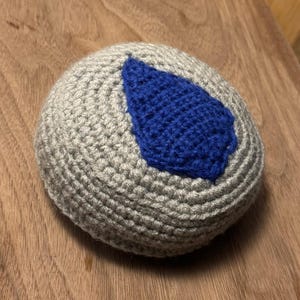 May include: A crocheted ball with a gray base and a blue leaf-shaped accent. The ball is round and appears to be made of soft yarn. The blue accent is a contrasting color and adds a decorative element.