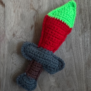 Può includere: Una spada all'uncinetto fatta a mano con punta verde, corpo rosso, guardia grigia e manico marrone. La spada giocattolo è realizzata in filato ed è progettata per il gioco dei bambini.