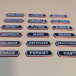 Könnte beinhalten: Achtzehn kleine, sechseckige Spielmarker, angeordnet in sechs Reihen. Jeder Marker ist dunkelbraun mit einem weißen Rand und zeigt weißen, geprägten Text, darunter Double Strike, Lifelink, Goaded, Indestructible, Trample, First Strike, Infect, Flying, Shroud, Hexproof, Ward, Reach, Vigilance, Defender, Deathtouch, Menace, Persist und Haste.