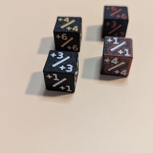 Könnte beinhalten: Drei schwarze sechsseitige Würfel mit texturierten Oberflächen und aufgedruckten Zahlenwerten. Jede Seite zeigt eine bruchartige Notation mit positiven Ganzzahlen, einschließlich Kombinationen wie +4/+4, +6/+6, +3/+3, +1/+1 und +6/+6 in weißer oder roter Tinte.