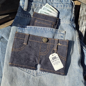 Può includere: Un portafoglio in denim scuro, a forma di pantaloncini di jeans, con cuciture gialle e un bottone, è appoggiato nella tasca di un paio di jeans azzurri. Il portafoglio contiene banconote. Un secondo portafoglio è visibile nella tasca posteriore.