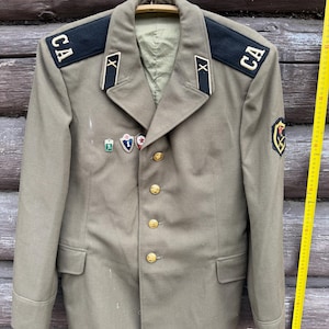 May include: Olive green military jacket with gold buttons, shoulder boards with "CA" lettering, and various insignia. The jacket is hanging on a wooden hanger against a weathered wood background. A yellow measuring tape is visible.