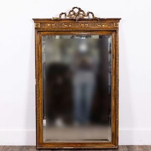 Peut inclure: Un miroir mural rectangulaire doté d'un cadre en bois foncé avec des accents dorés. Le haut du cadre est orné d'un motif de ruban noué sculpté et d'une frise décorative. La surface en verre est claire et réfléchissante avec un bord biseauté.